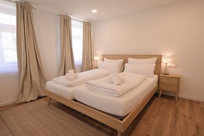 Una cama doble con ropa de cama y toallas blancas, cabecera de madera, lámparas de noche y una alfombra tejida sobre piso de madera.