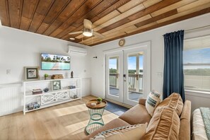 A living room with a wooden ceiling, a TV mounted on the wall, a white console table, a sofa, and a sliding glass door leading to a balcony. - Birdwatch Bungalow Upstairs (Cedar Key)