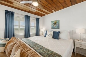A bedroom with a large bed, a bedside table with a lamp, a wall-mounted painting, and a window with curtains. - Birdwatch Bungalow Upstairs (Cedar Key)