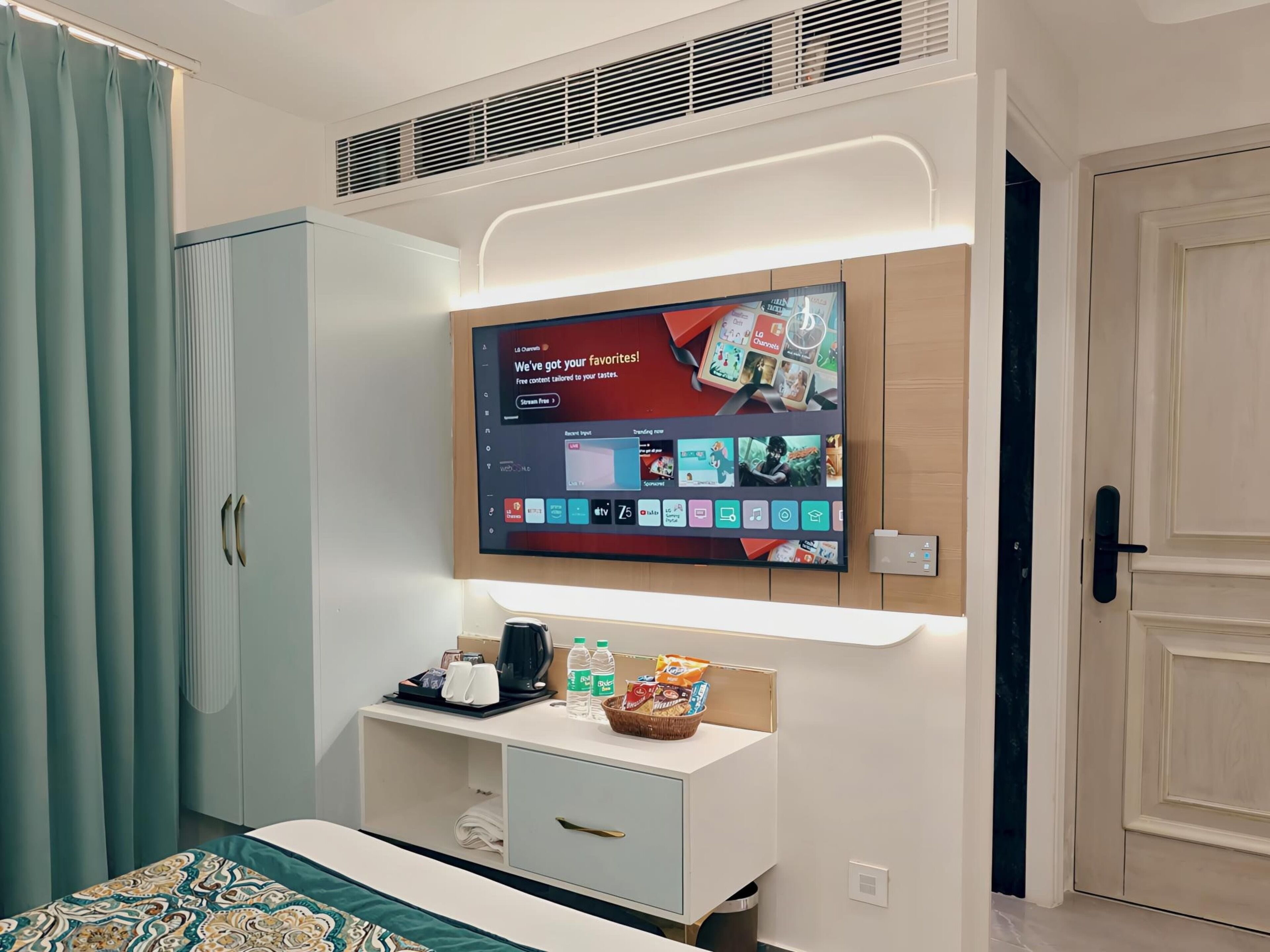 Una habitación de hotel moderna con un televisor de pantalla plana montado en la pared, una cama con ropa de cama estampada, una mesita de noche con una tetera, tazas y aperitivos, y un armario con un espejo.