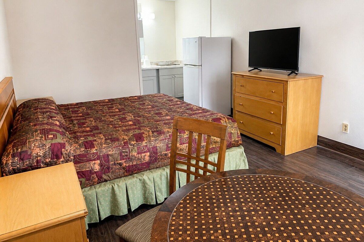 A bedroom with a bed, a wooden dresser, a television, and a kitchenette in the background.