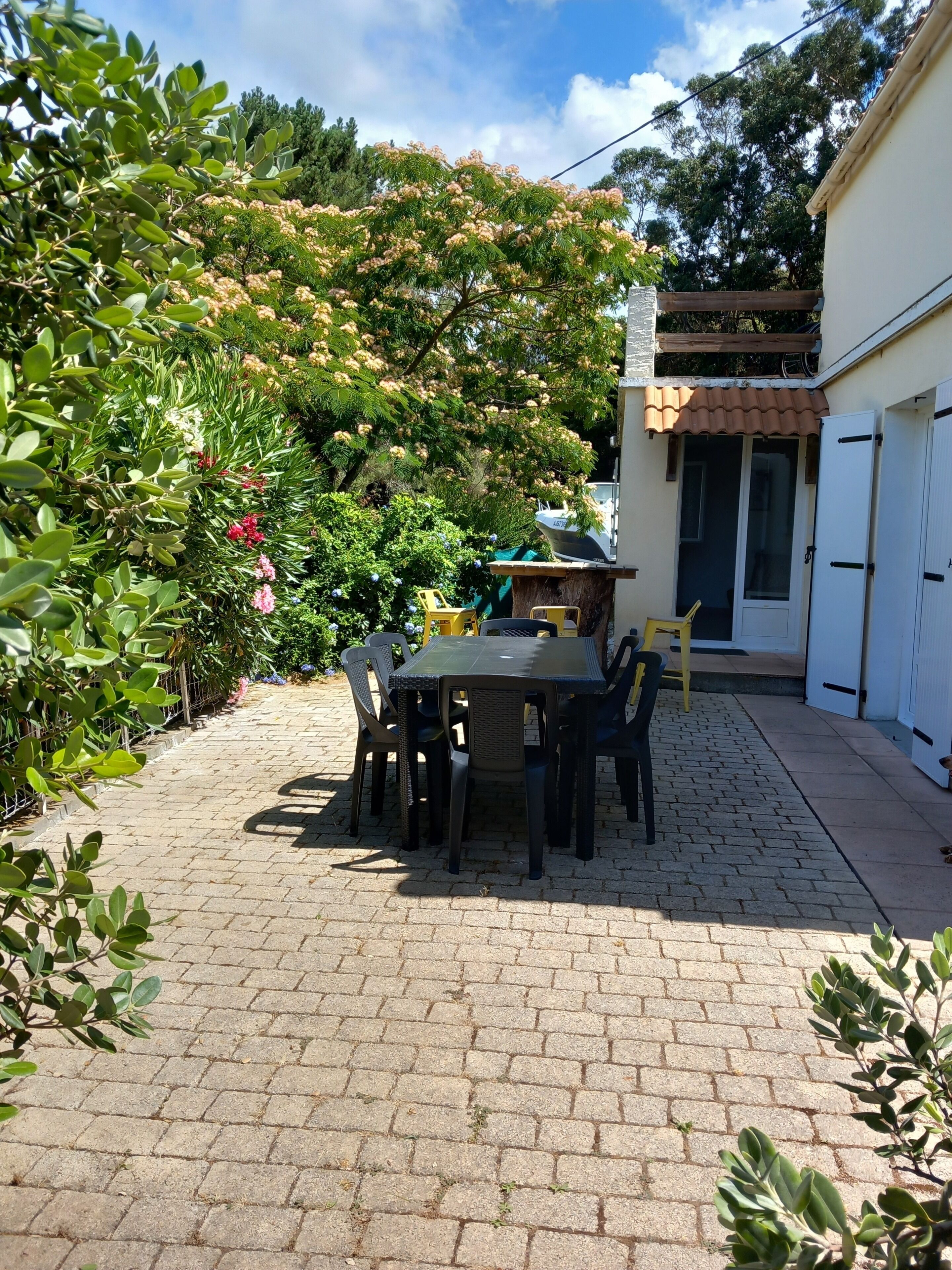 A patio with a table and chairs, surrounded by greenery and a building with a tiled roof.