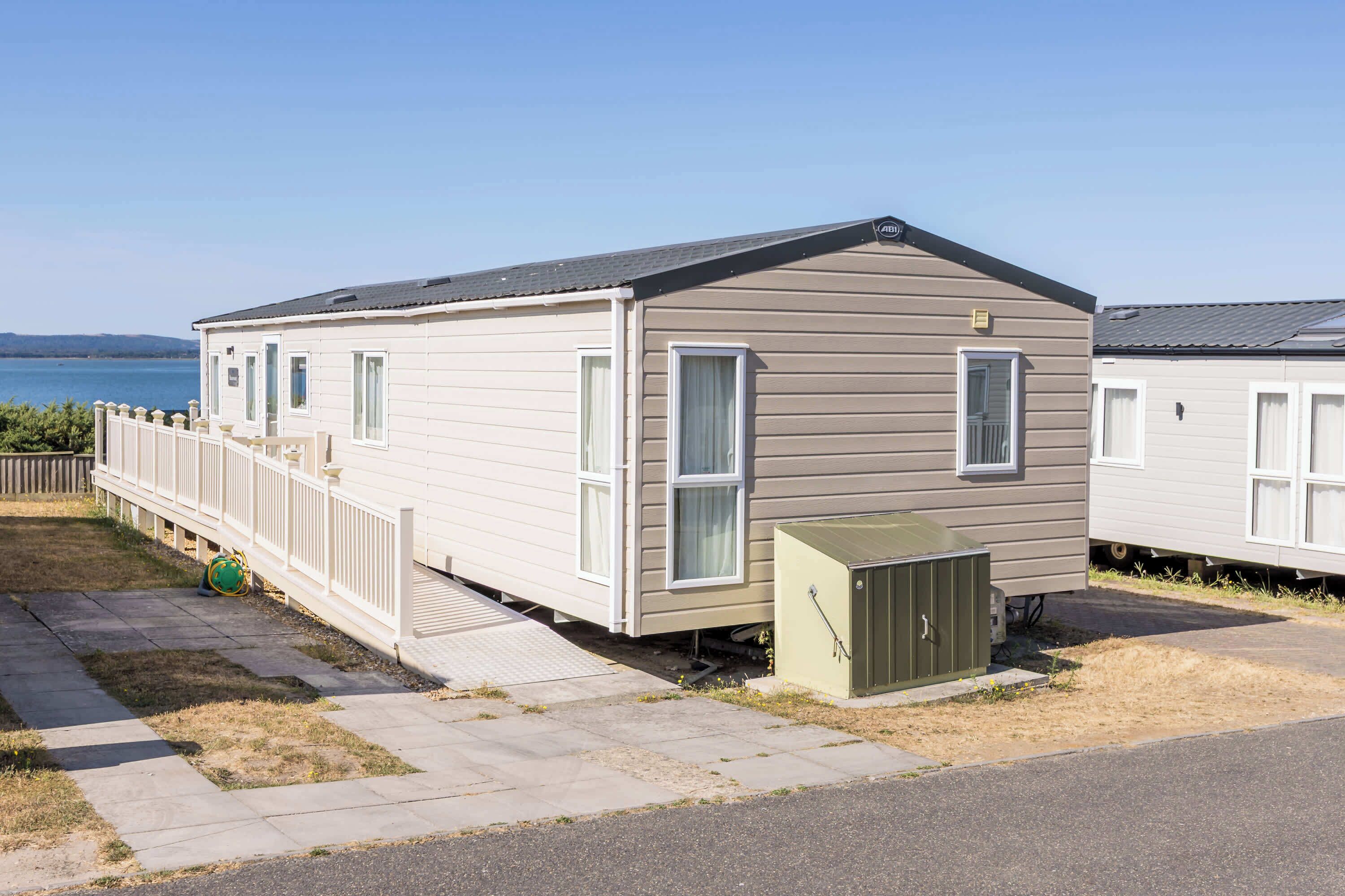 Une maison mobile de plain-pied dotée d’une terrasse clôturée, d’un petit abri de jardin attenant et offrant une vue dégagée sur le plan d’eau en arrière-plan.
