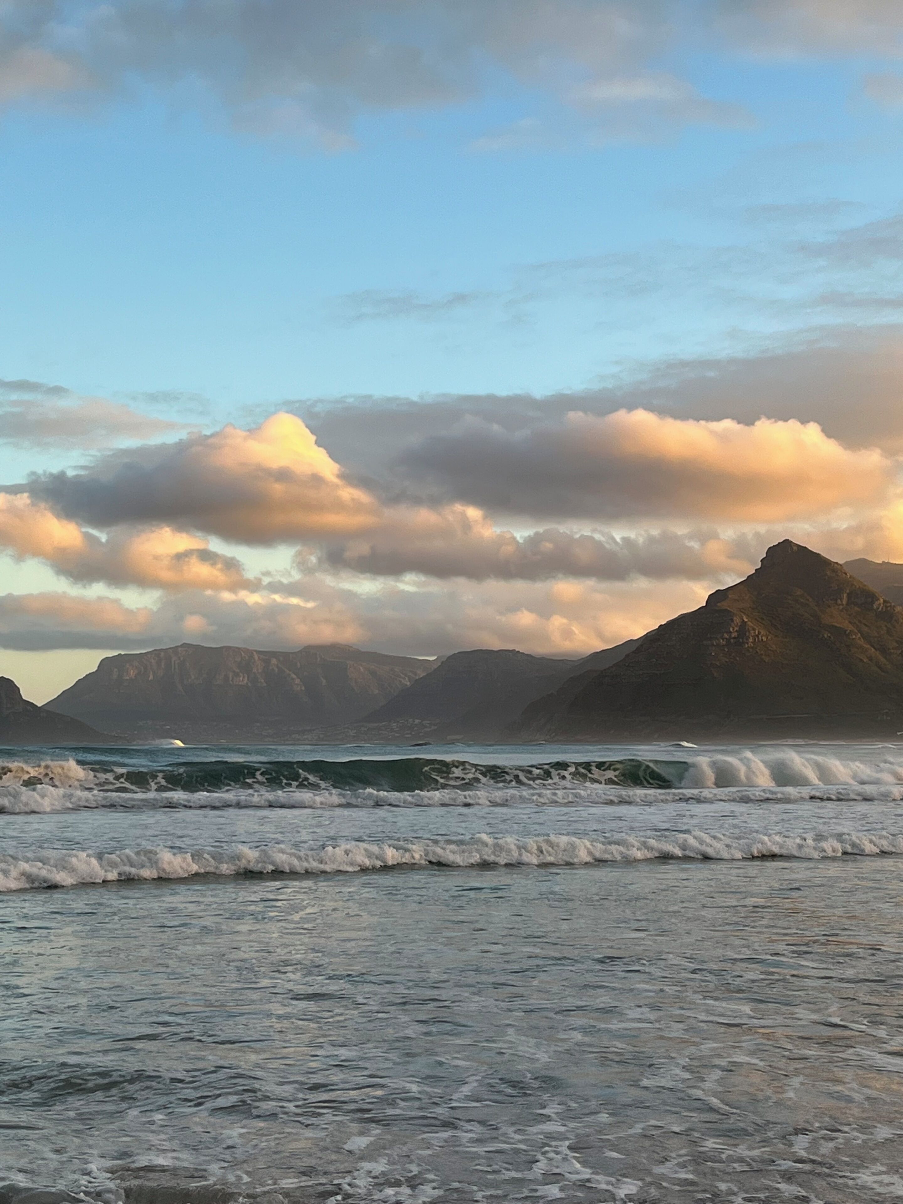 A coastal landscape with waves, mountains, and a partly cloudy sky.