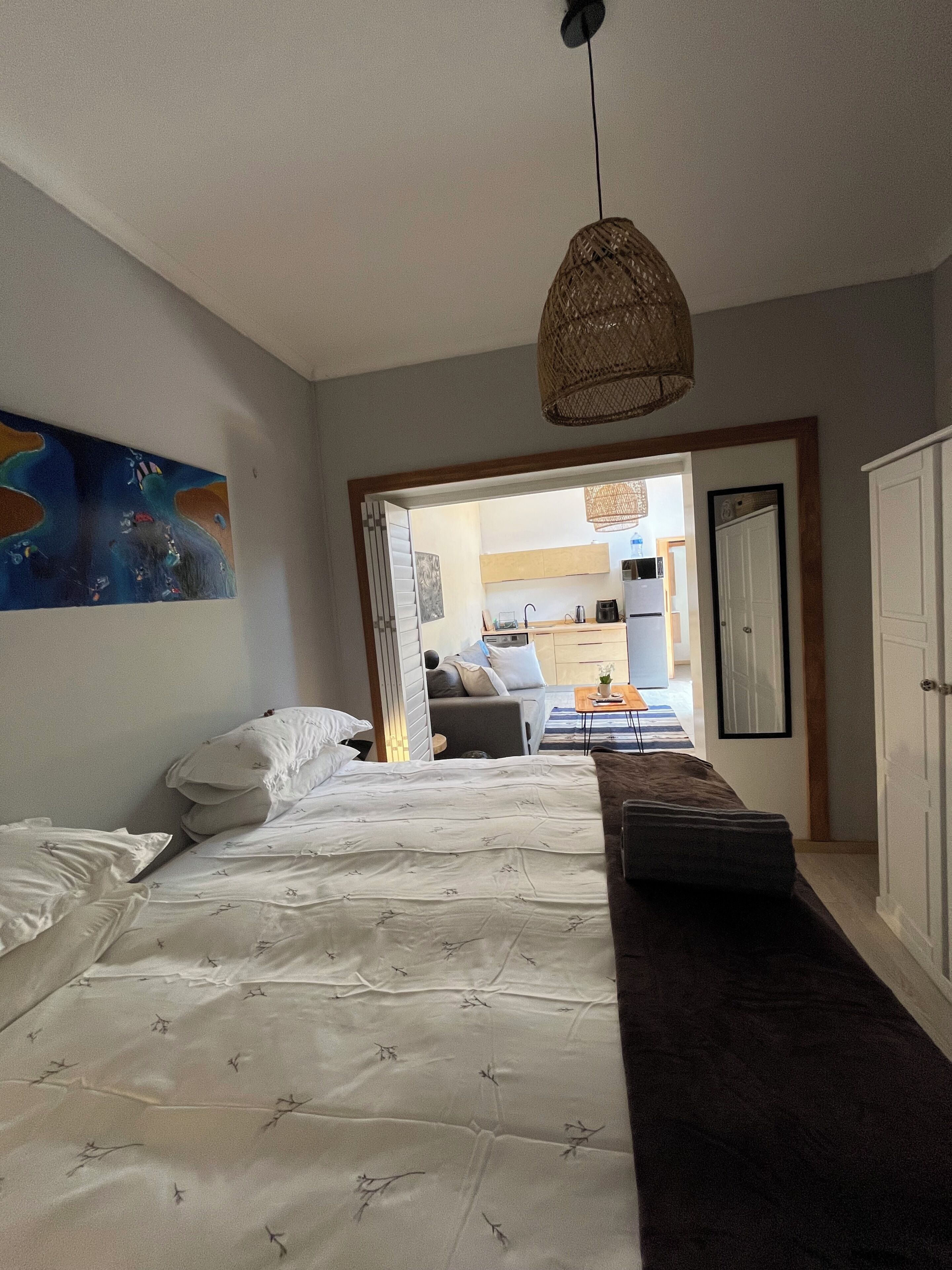 A bedroom with a bed, a hanging light fixture, a painting on the wall, and a view into the adjacent room.