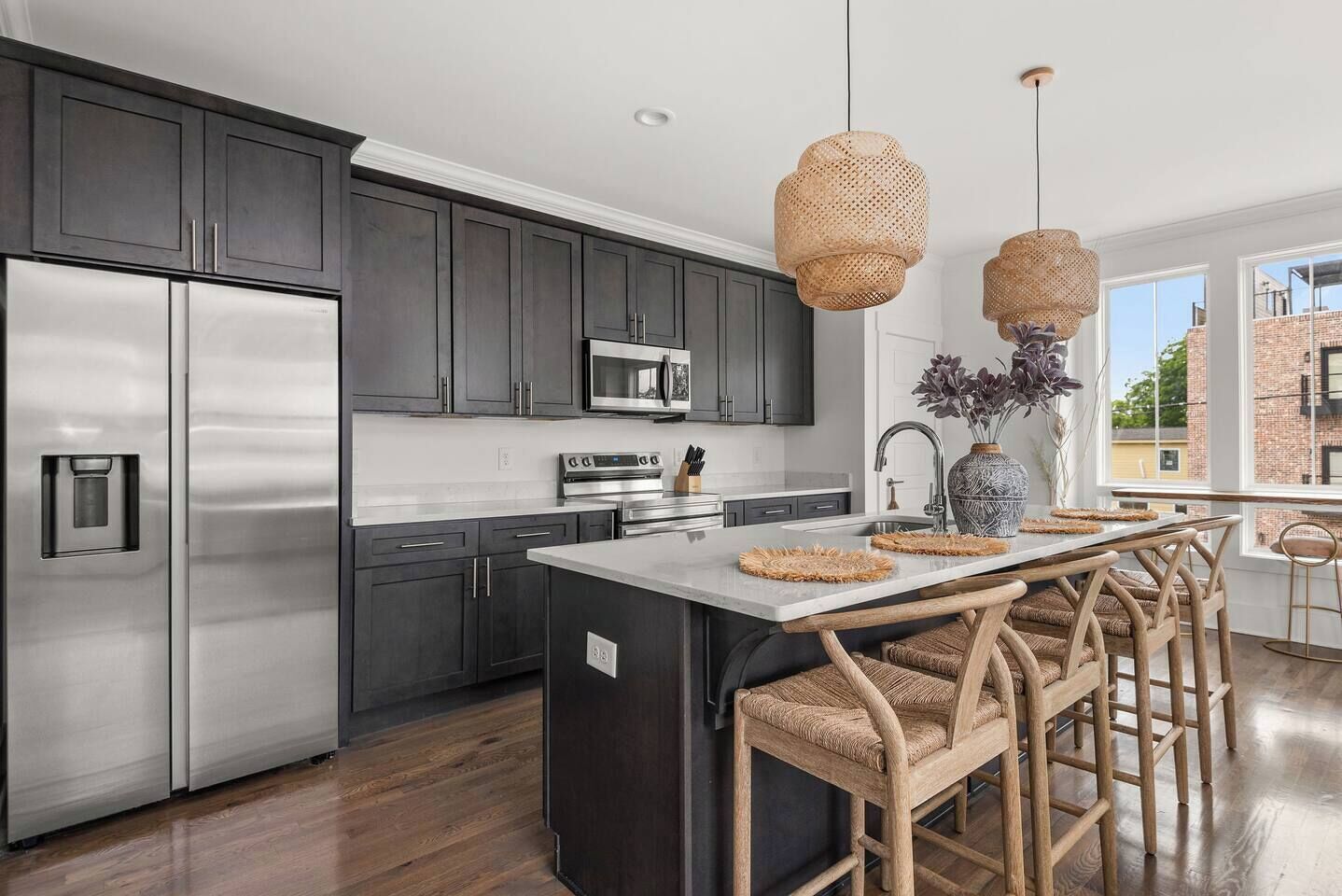 A modern kitchen with dark cabinets, stainless appliances, a central island with bar stools, and a large window offering a view.