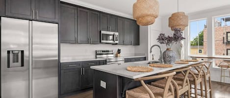 A modern kitchen with dark cabinets, stainless appliances, a central island with bar stools, and a large window offering a view.