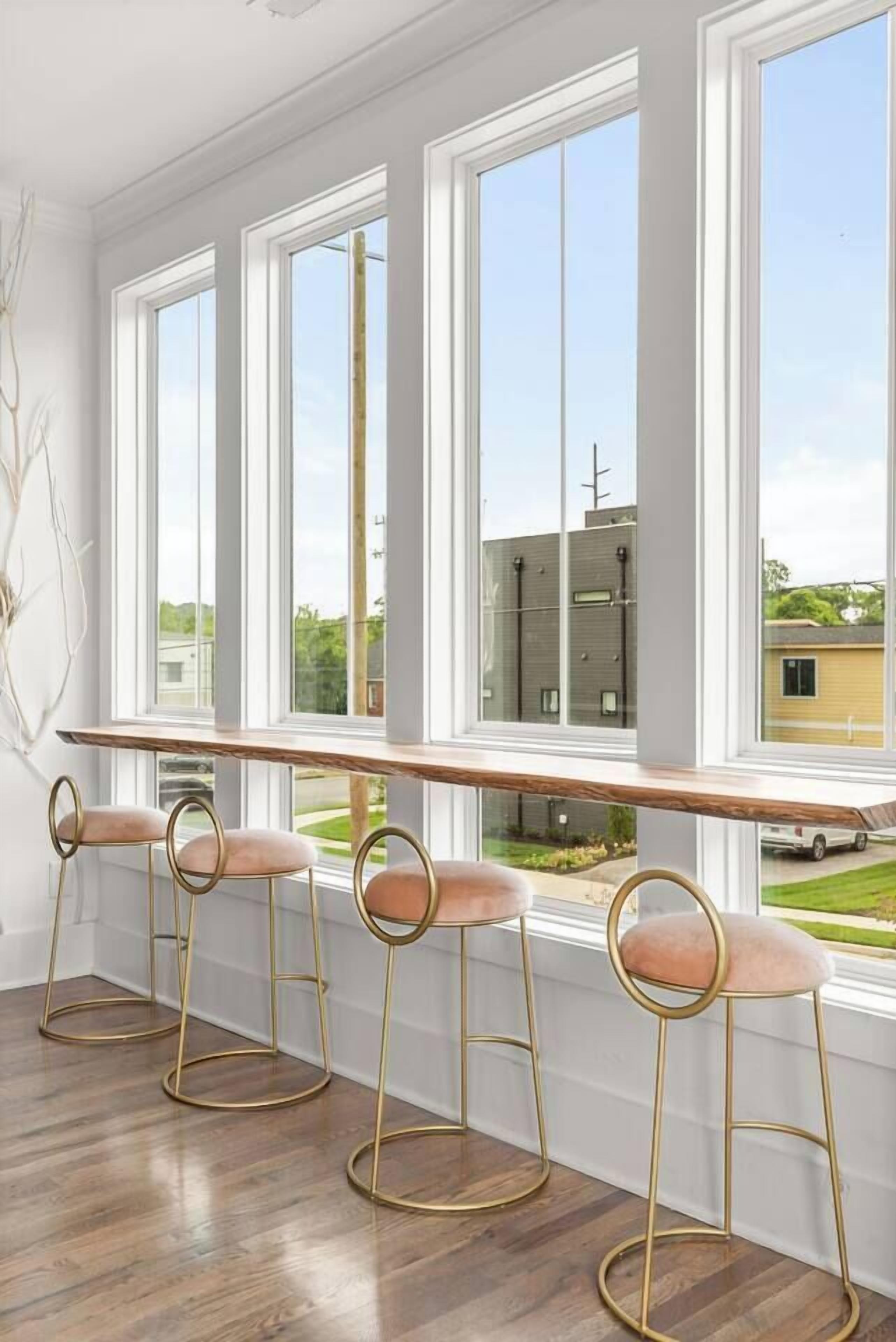 A room with a long window seat with a wooden ledge, three bar stools with cushioned seats, and a view of a building and greenery outside.