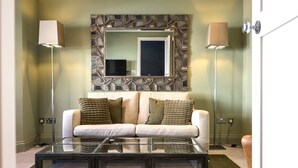 Ein modernes Wohnzimmer mit einem Glastisch, einer Couch mit Kissen, einem Wandspiegel und zwei Stehlampen.