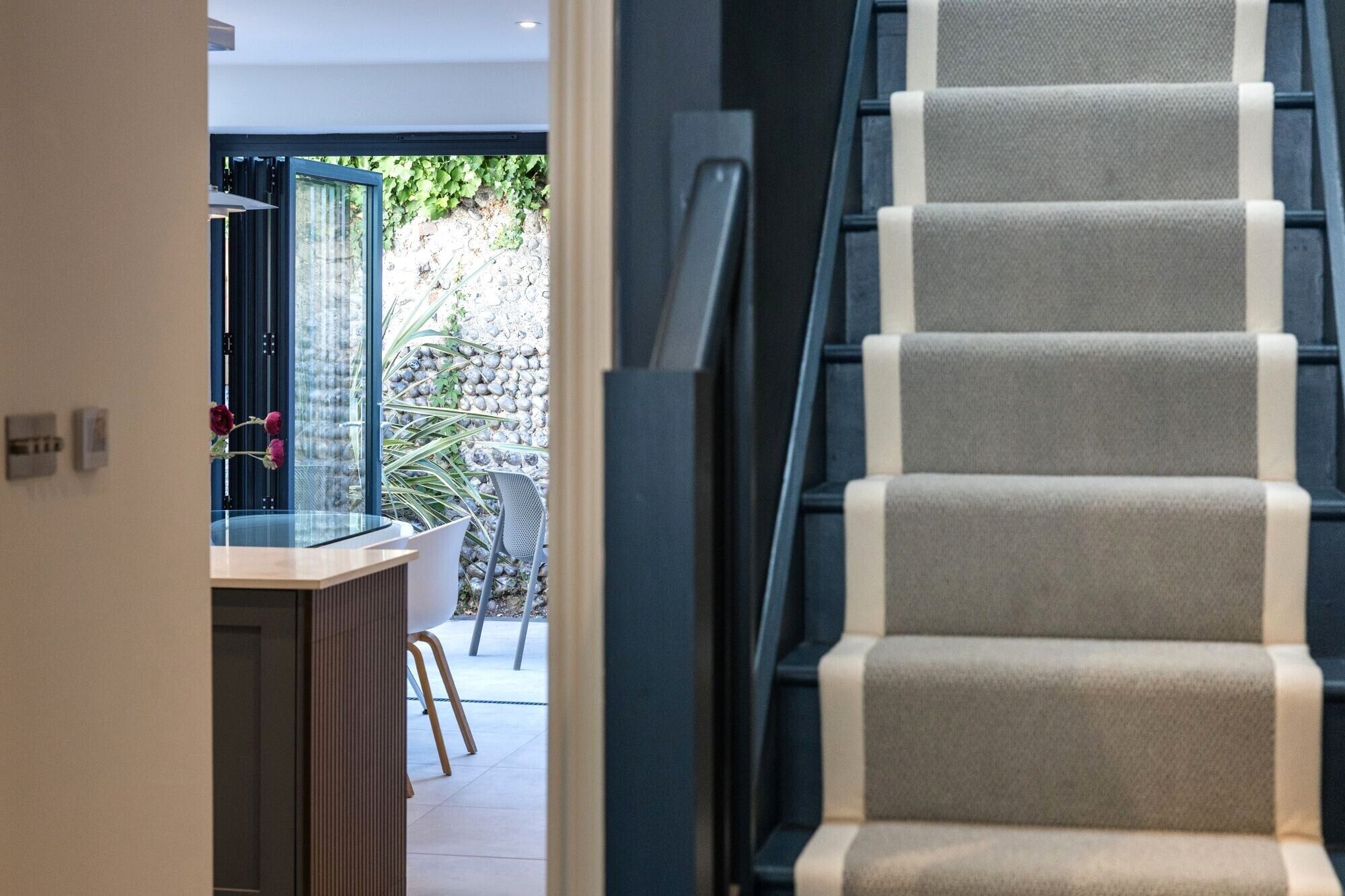 A staircase with a modern design, leading to an outdoor area with a stone wall and greenery.