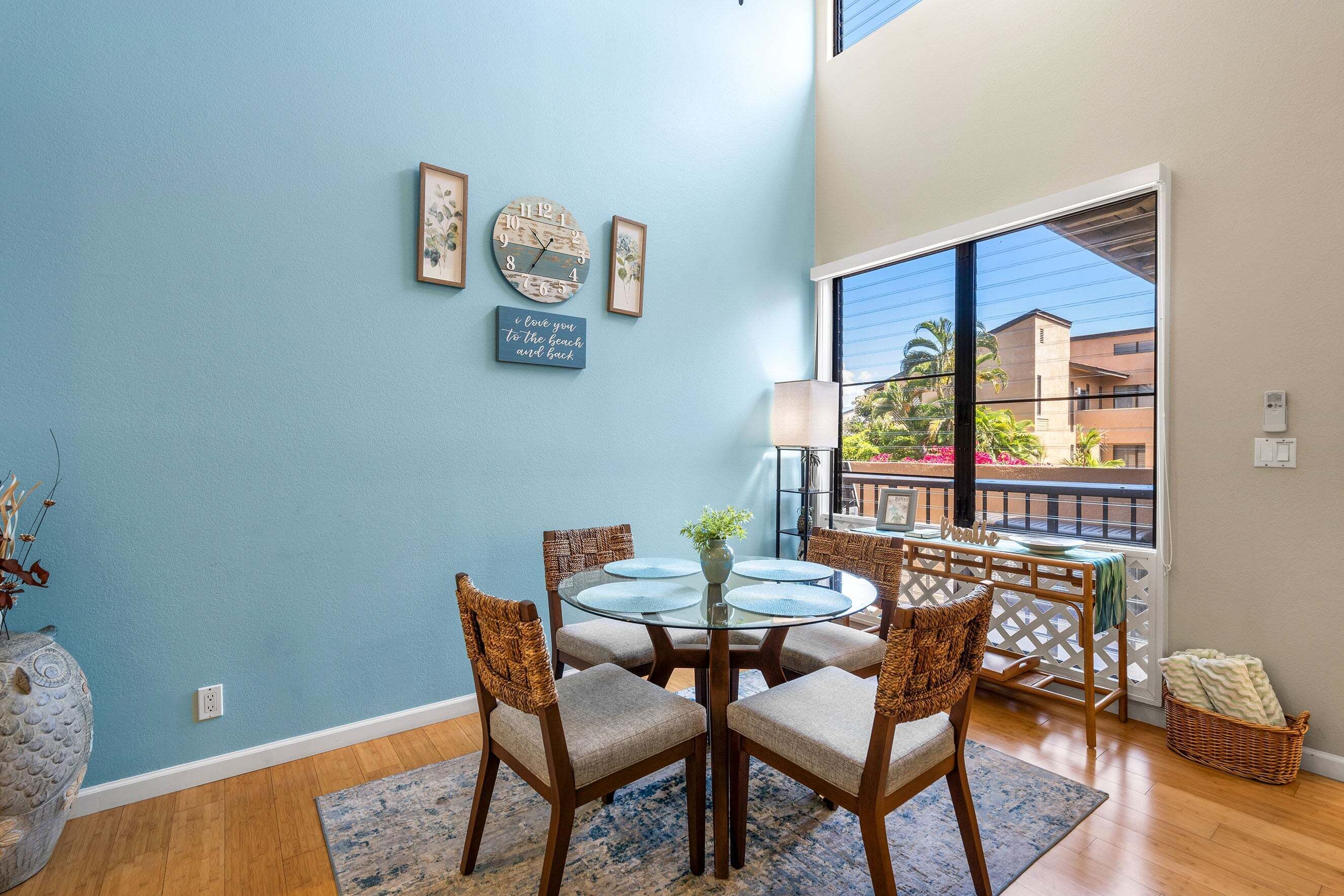 A dining area with a round glass table, wicker chairs, a blue wall, and a view of buildings through a large window.