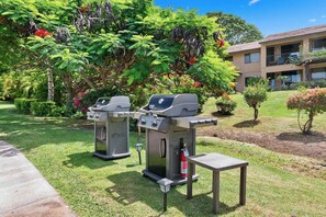 Two barbecue grills, a small table, and a fire extinguisher in a grassy area with a building in the background. - Wailea Ekahi 52D - Wailea 1BR Condo, Garden Lanai, AC, Island Luxury Beach Condo (Kihei)
