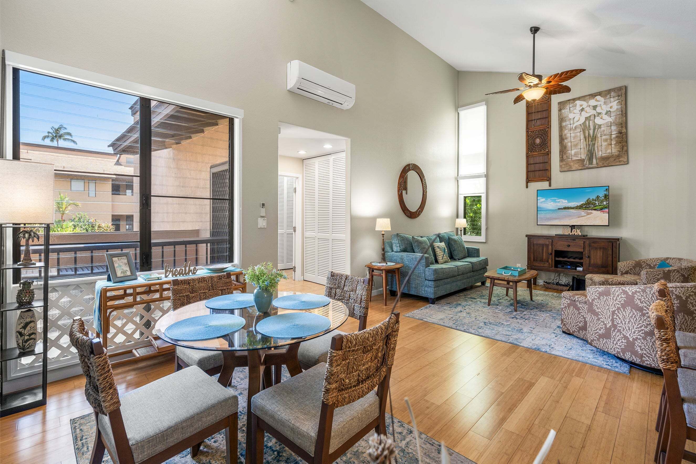 A spacious living room with a dining area, a ceiling fan, a TV, and a large window overlooking a balcony.