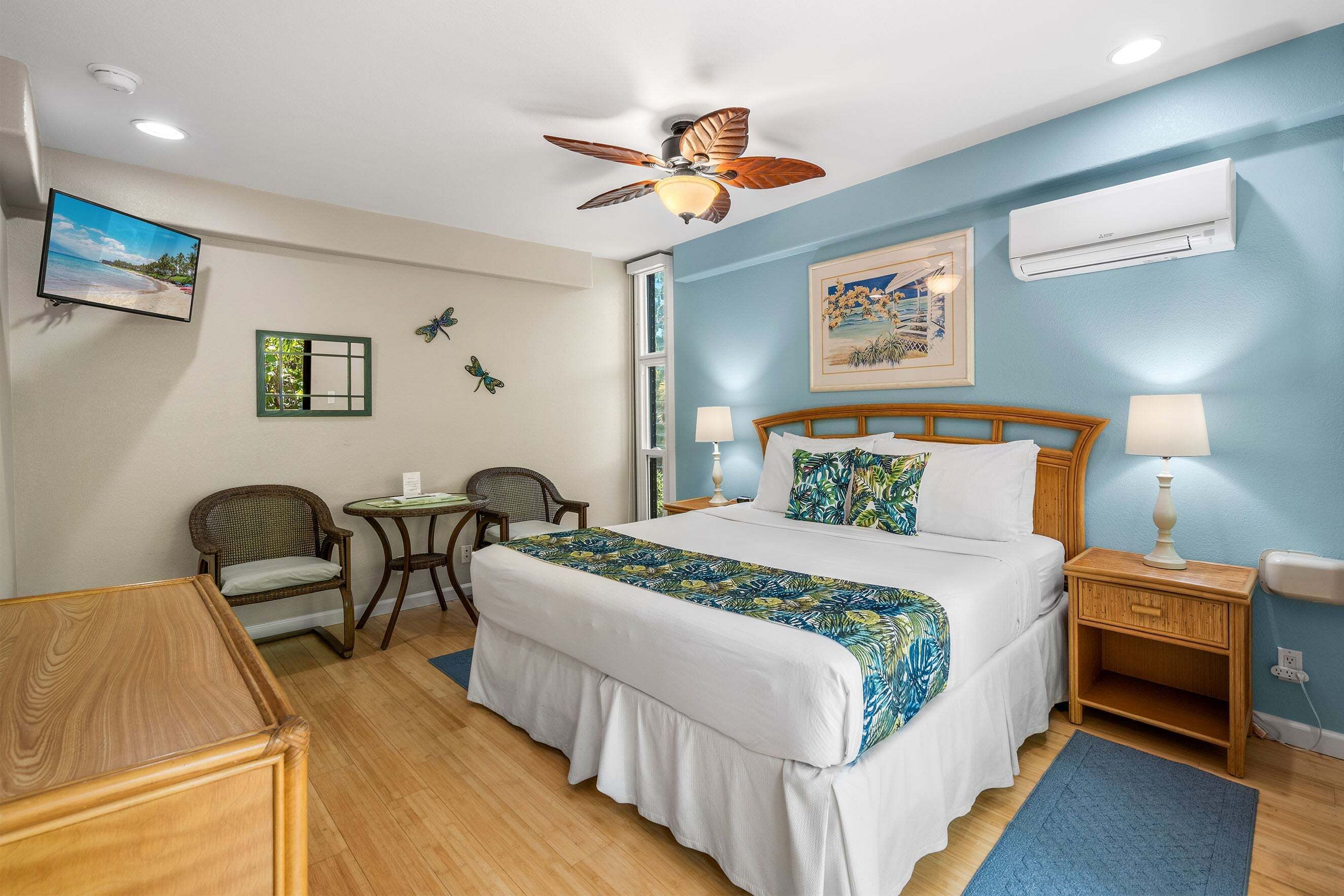 A hotel room with a bed, a ceiling fan, a TV, a chair, a table, and an air conditioning unit.