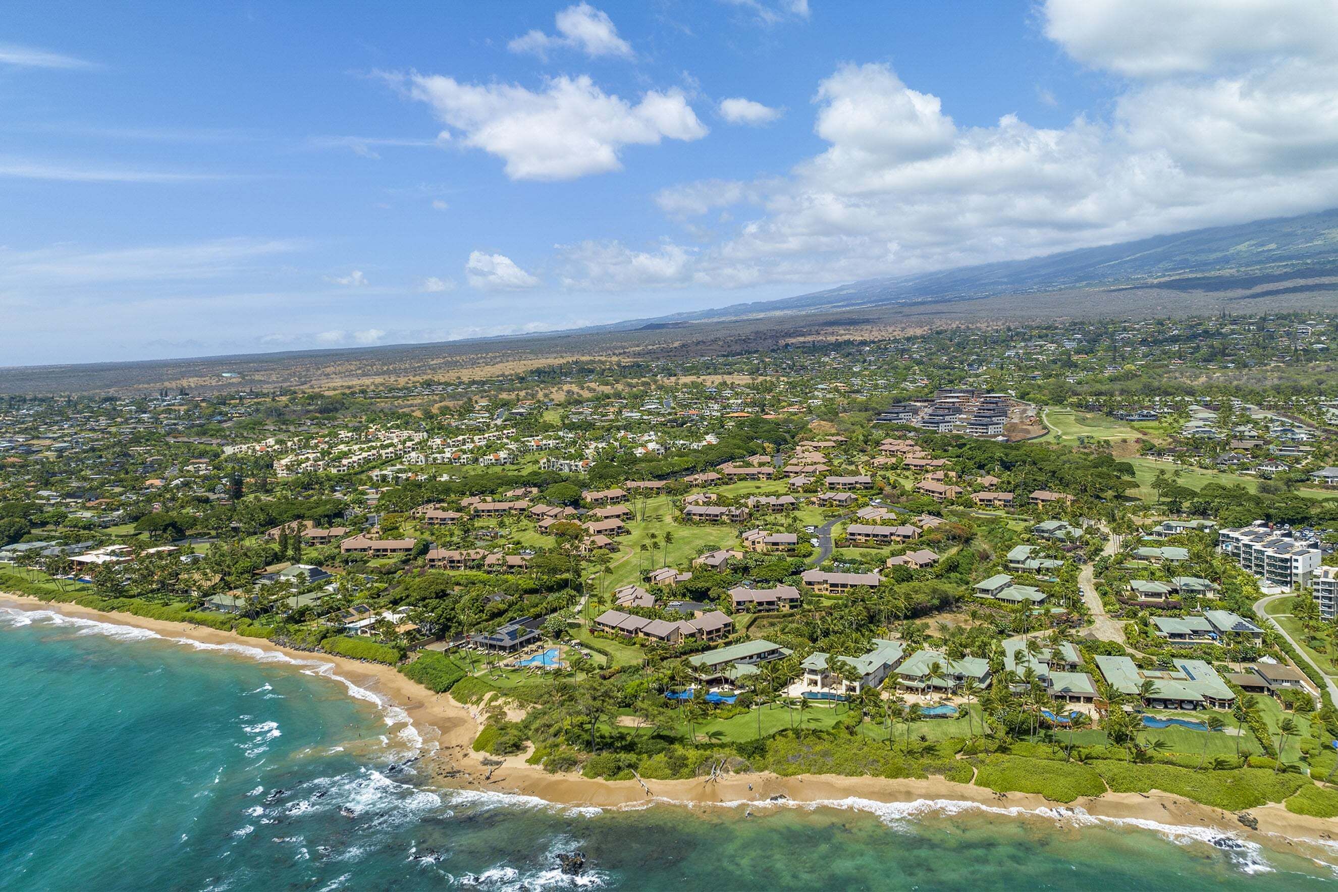 An aerial view of a coastal resort with multiple buildings, a beach, and surrounding greenery.