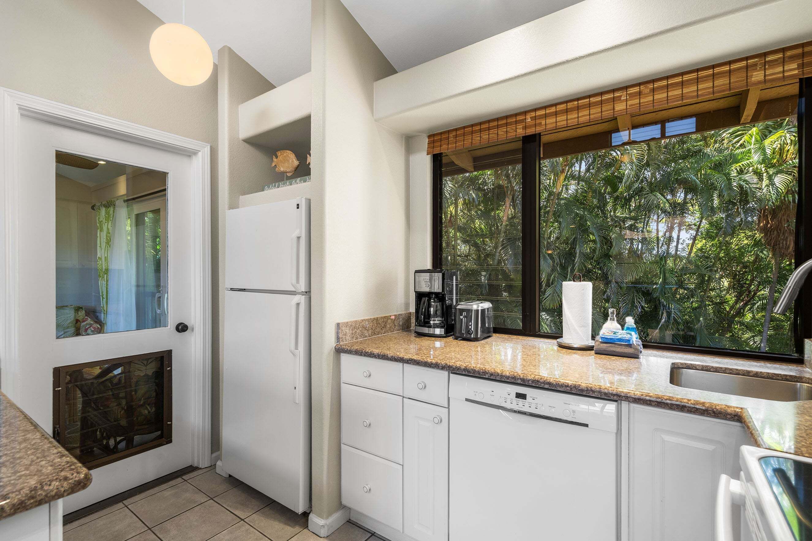 A kitchen with a window view of greenery, a coffee maker, and a refrigerator.