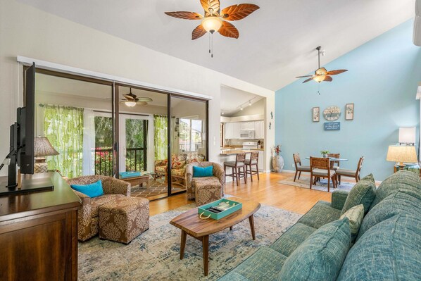 A spacious living area with a sofa, coffee table, and a dining table with chairs. There is a ceiling fan, a kitchen in the background, and a sliding glass door leading to a balcony. - Wailea Ekahi 52D - Wailea 1BR Condo, Garden Lanai, AC, Island Luxury Beach Condo (Kihei)