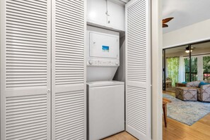 Habitación compacta con lavadora y secadora integradas, una mesita y vistas a un espacio exterior con un sofá y cortinas.