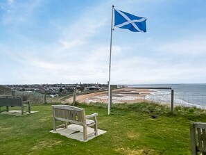 Eine grüne Fläche mit einem Fahnenmast, einer Bank und Blick auf das Meer sowie entfernte Gebäude.