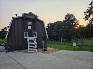 Ein hölzernes Bauernhaus mit einer Leiter, die zu einem Eingang auf den Dachboden führt, umgeben von einer Wiese und Bäumen.