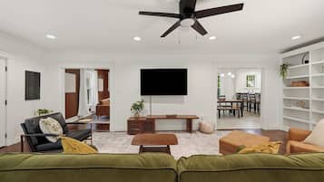 A modern living room with a ceiling fan, a flat-screen TV, and a dining area visible through an open door.