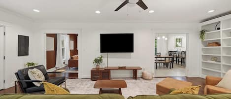Un salon moderne doté d’un ventilateur de plafond, d’un téléviseur à écran plat et d’un coin repas visible à travers une porte ouverte.