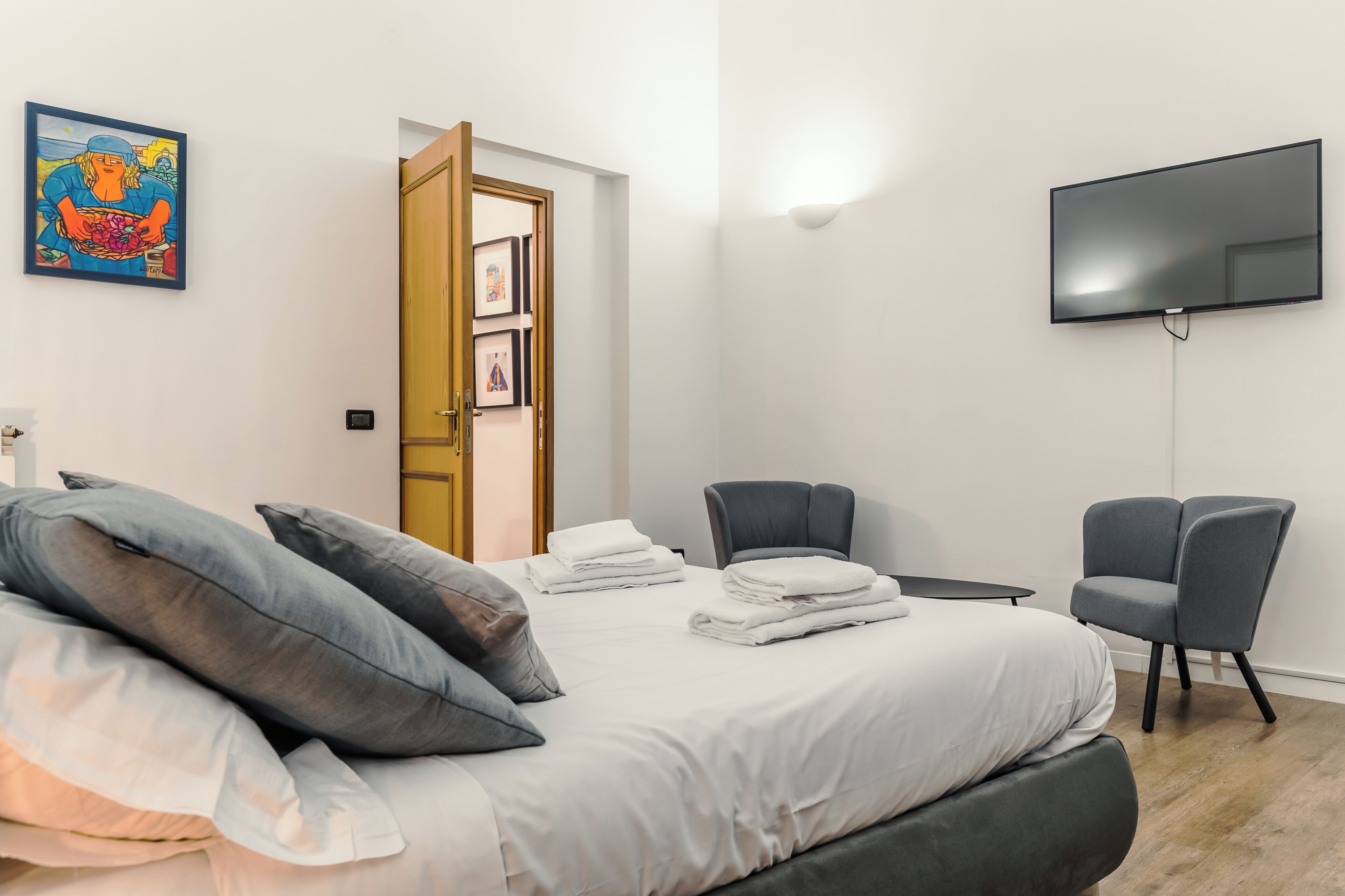 Habitación de hotel con cama, dos sillas, televisión de pantalla plana y puerta que da a un pasillo.