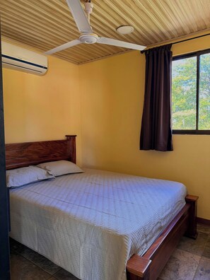 Ein Schlafzimmer mit Bett, Holz-Kopfstück, Deckenventilator, Klimaanlage, Fenster mit Vorhängen und Blick ins Grüne.
