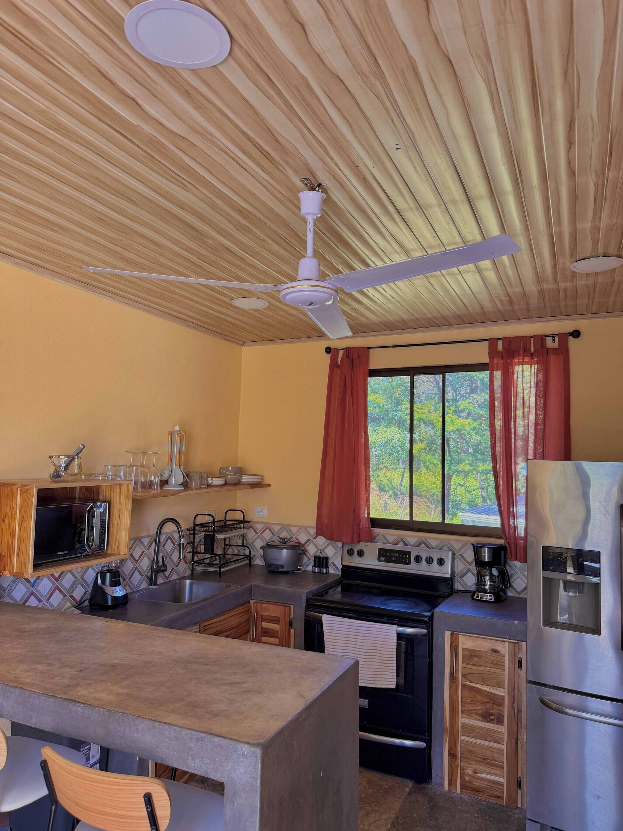 A kitchen with a wooden ceiling, a stove, a refrigerator, and a window with curtains.
