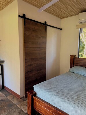 A bedroom with a wooden bed frame, a pull-down shutter door, and a window with an air conditioning unit. - Cueva Small Apartment Saltwater Pool Hilltop View (Cabo Velas)