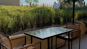 Eine Terrasse im Freien mit einem Glastisch und Stühlen, umgeben von Grünpflanzen und einem Gebäude im Hintergrund.