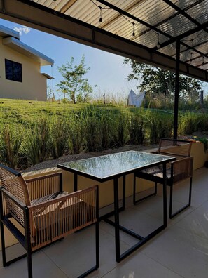 An outdoor patio with a glass-top table and chairs, surrounded by greenery and a building in the background. - Cueva Small Apartment Saltwater Pool Hilltop View (Cabo Velas)