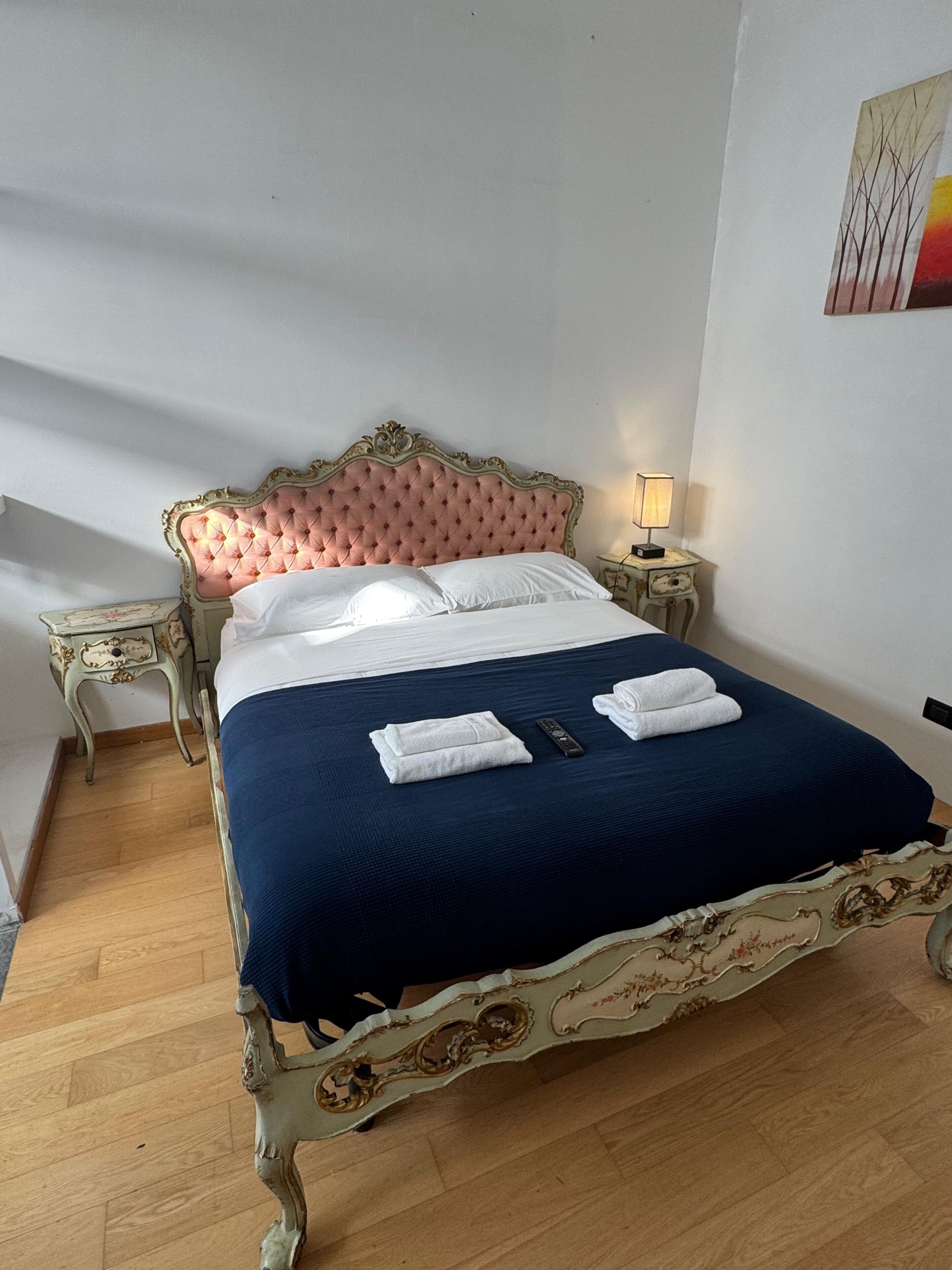 A bed with a tufted headboard, a blue bedspread, and two folded towels. There is a nightstand with a lamp and a small table with a decorative item.