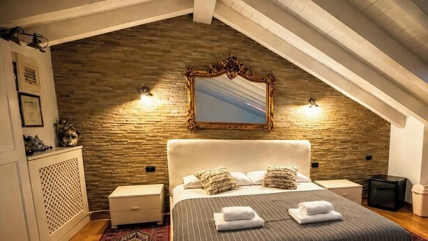 Un dormitorio con pared de piedra, una cama grande con almohadas, una mesita de noche, un aparador y un espejo.