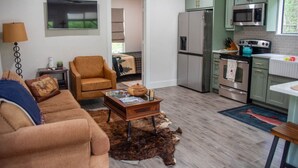 Ein Wohnzimmer mit einer Couch, einem Couchtisch und einem Sessel. Es gibt eine Küchenzeile mit Kühlschrank, Herd und Mikrowelle.