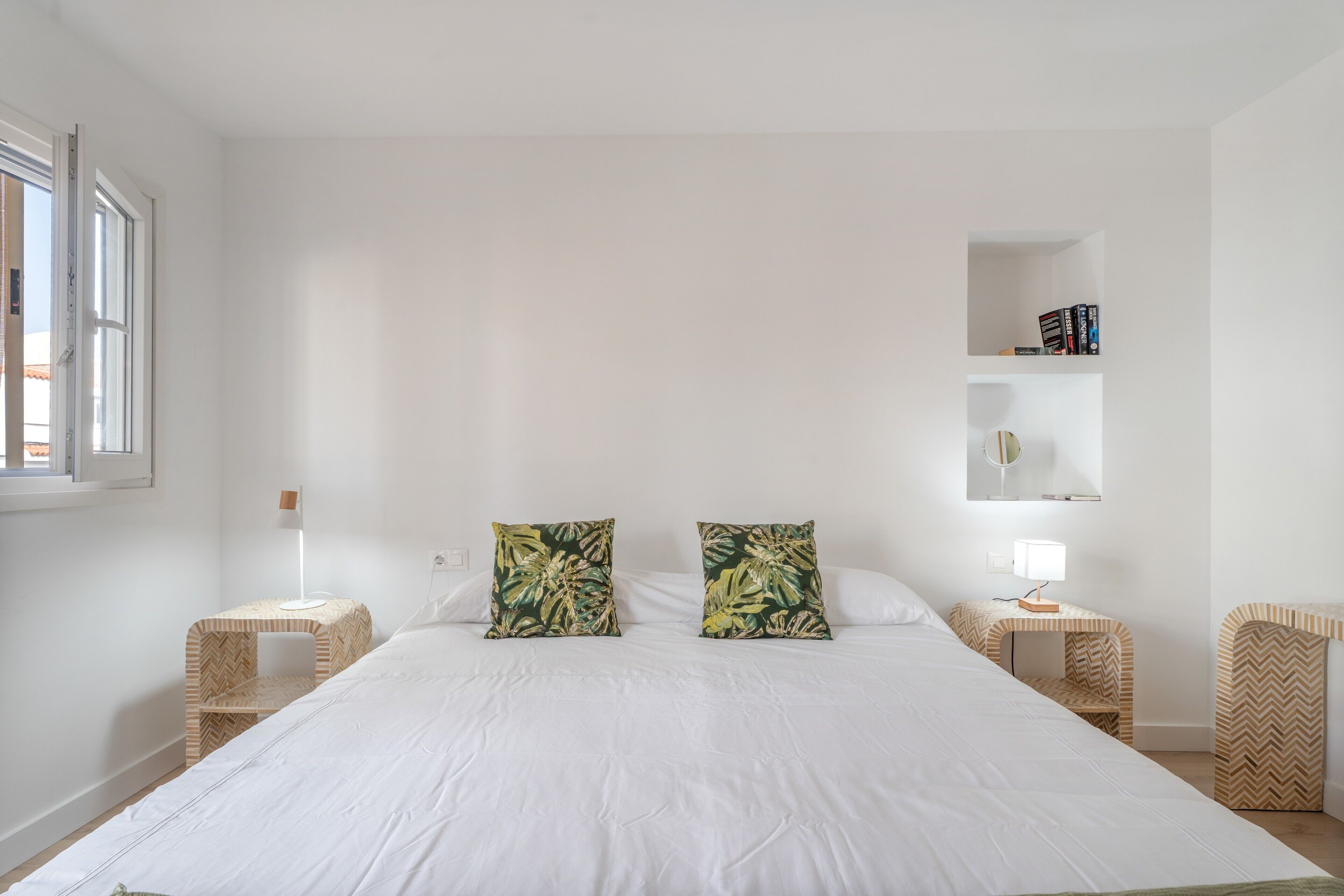 Een minimalistische slaapkamer met een groot bed, twee nachtkastjes, een bijzettafeltje, een plank met boeken en een raam.