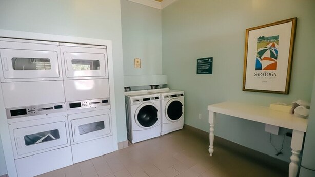Une buanderie équipée d’une machine à laver et d’un sèche-linge superposés, d’une table avec une lampe et d’un tableau encadré au mur.