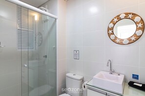 Ein Badezimmer mit Duschkabine aus Glas, Toilette, Waschbecken mit Spiegel und einem an der Wand befestigten Toilettenpapierhalter.