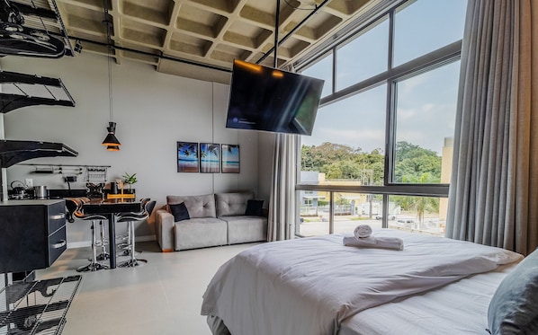 A modern hotel room with a large bed, a desk with a chair, a sofa, and a TV mounted on the wall. - Nice Comfort 750m From Canas Beach Tps203 (Florianópolis)