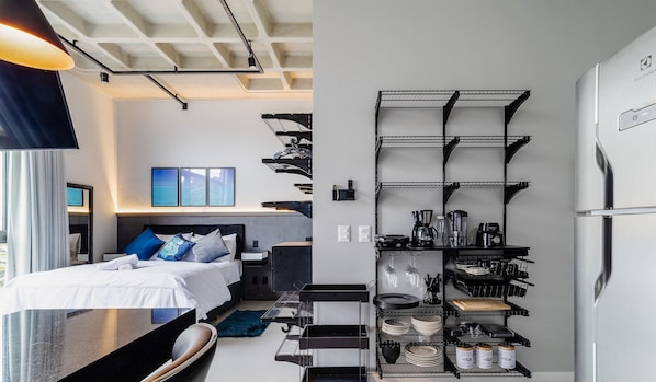 A modern hotel room with a bed, bedside table, and a built-in kitchen area with shelves and appliances. - Nice Comfort 750m From Canas Beach Tps203 (Florianópolis)