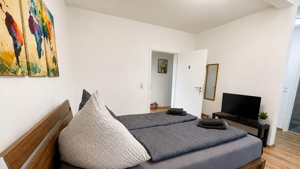 Um quarto com uma cama, uma televisão e uma porta que dá para outro cômodo.