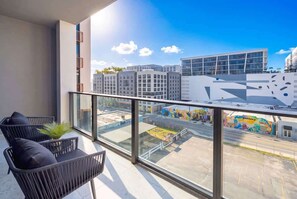 A balcony with two chairs and a potted plant, overlooking a cityscape with modern buildings and a mural. - Luxury Wynwood Studio with Pool, Gym & NoMad Bar (Miami)