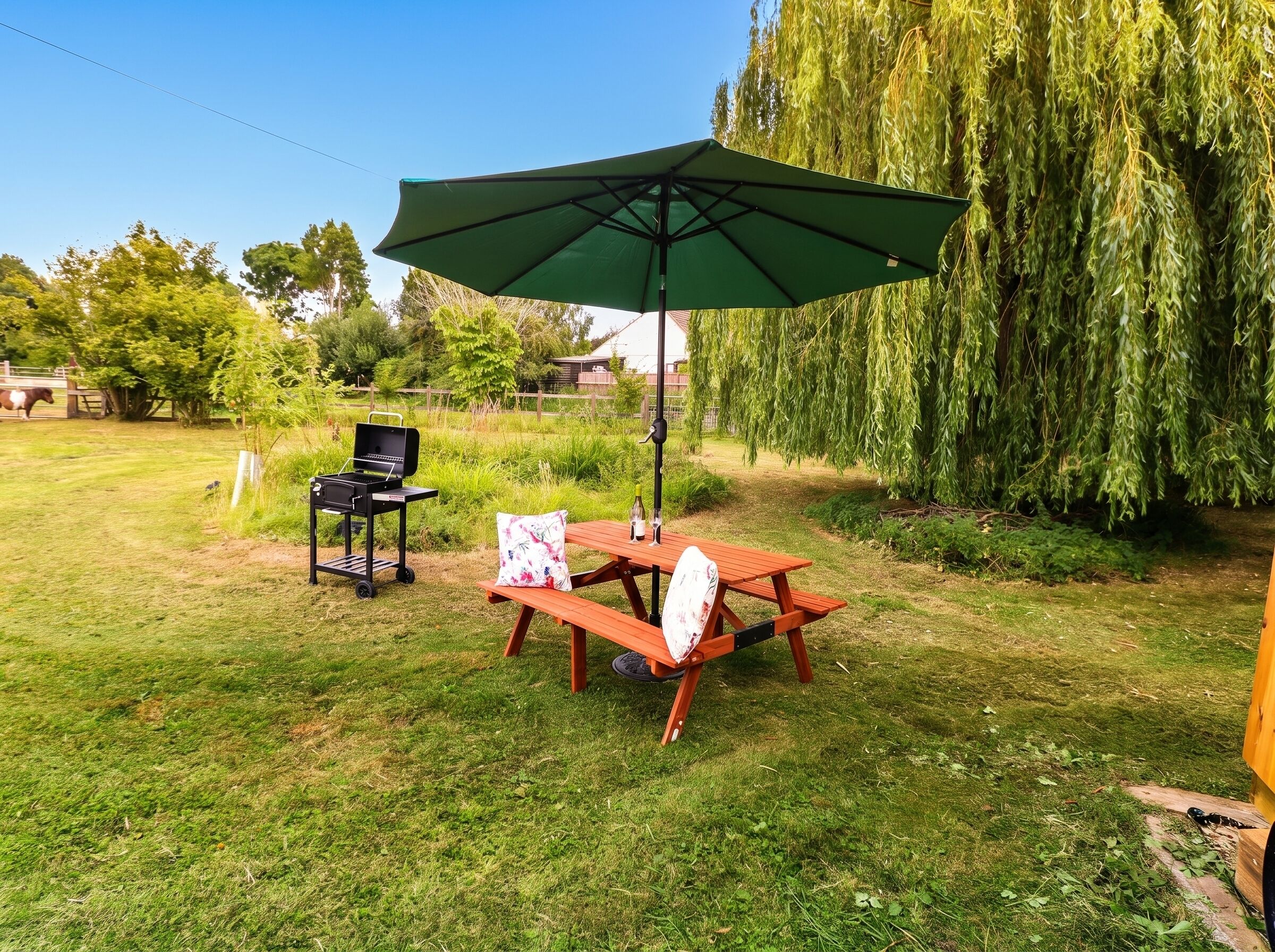 An outdoor patio area with a wooden table and benches, a barbecue grill, and a green umbrella providing shade.