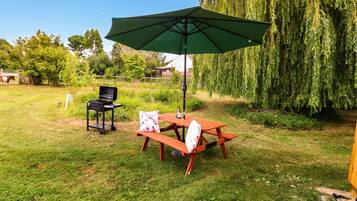 An outdoor patio area with a wooden table and benches, a barbecue grill, and a green umbrella providing shade.