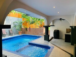 A swimming pool with a view of tropical foliage, a ceiling fan, and a small kitchen area.