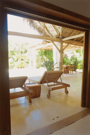 A covered outdoor seating area with wooden lounge chairs and a table, surrounded by greenery and a thatched roof structure. - 2-bedroom apartment in soothing Punta Cana with AC and gym access (Punta Cana)