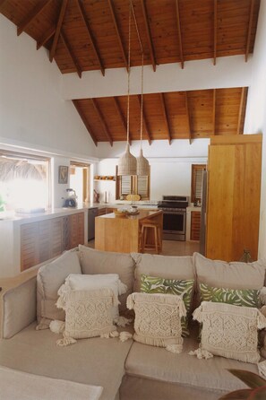 A spacious living area with a high wooden ceiling, a kitchen in the background, and a seating area with macramé cushions. - 2-bedroom apartment in soothing Punta Cana with AC and gym access (Punta Cana)