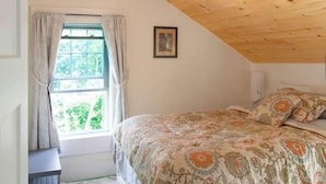A bedroom with a sloped ceiling, a bed with patterned bedding, a window with curtains, and a door with a light switch. - Lock`s Nest on Peaks Island Maine (Portland)