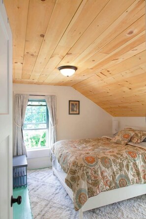 Ein Schlafzimmer mit schrägem Dach, einem Bett mit gemusterten Betttextilien, einem Fenster mit Vorhängen und einer Tür mit Lichtschalter.