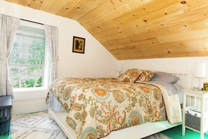A bedroom with a wooden ceiling, a bed with patterned bedding, a window with curtains, and a bedside table with a lamp. - Lock`s Nest on Peaks Island Maine (Portland)